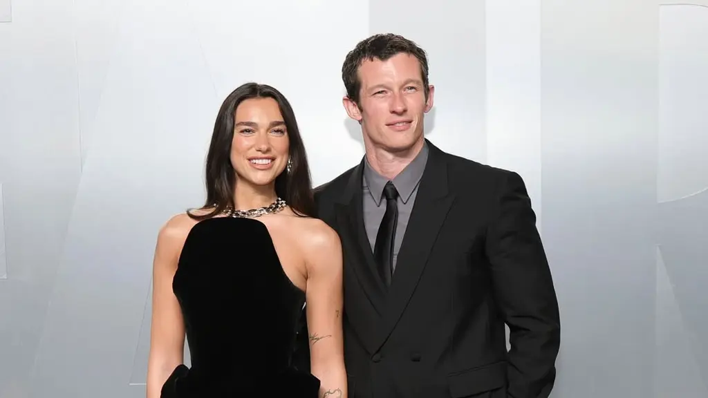 Dua Lipa tampil memukau dalam balutan gaun Schiaparelli bernuansa emas metalik dengan detail fur dramatis, menciptakan siluet sensual sekaligus bold. Ia tampak serasi dengan sang tunangan, Callum Turner, yang memilih setelan klasik bernuansa krem dengan bow tie hitam. Perpaduan mereka memancarkan aura old Hollywood glamour yang modern.