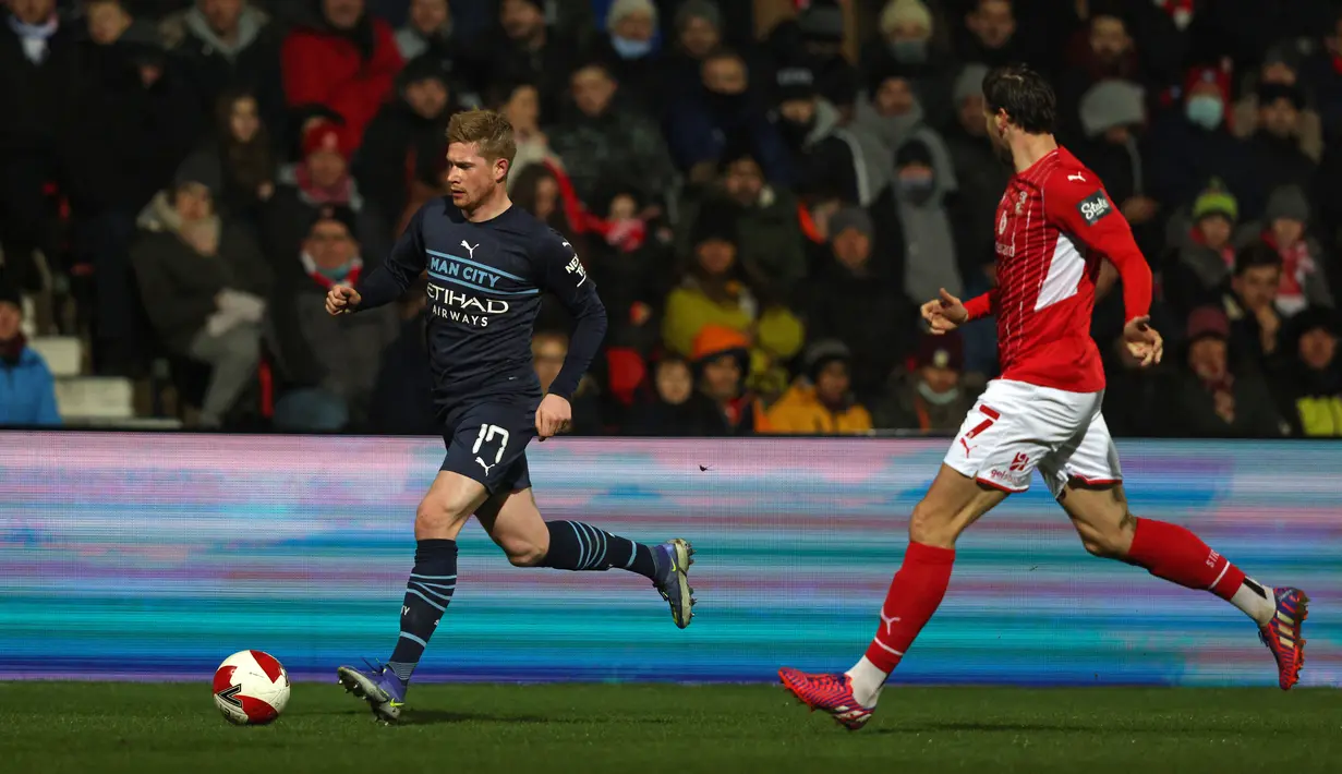 Gelandang Manchester City Kevin De Bruyne (kiri) berebut bola dengan gelandang tim divisi 4 Swindon Town, Ben Gladwin dalam duel putaran ketiga Piala FA 2021/22 di County Ground, Sabtu (8/1/2022) dini hari WIB. Manchester City menang telak 4-1 saat menghadapi Swindon Town. (Adrian DENNIS / AFP)