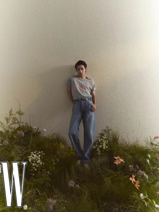 V BTS tampil kasual di antara daun dan bunga. Ia mengenakan kaus abu-abu dengan corak tulisan CELINE, dipadunya mengenakan celana jeans. [Foto: Instagram/wkorea]