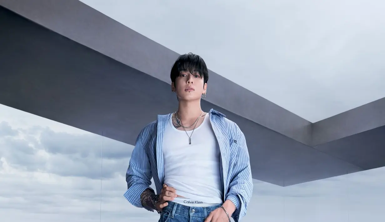 Nuansa berbeda hadir lewat tampilan smart casual. Jung Kook mengenakan kemeja striped biru yang dibuka santai di atas tank top putih dan jeans wide-leg. Siluet longgar memberikan kesan effortless sophistication, sementara latar interior modern dengan pemandangan laut memperkuat aura mature dan tenang. [Dok/Calvin Klein].