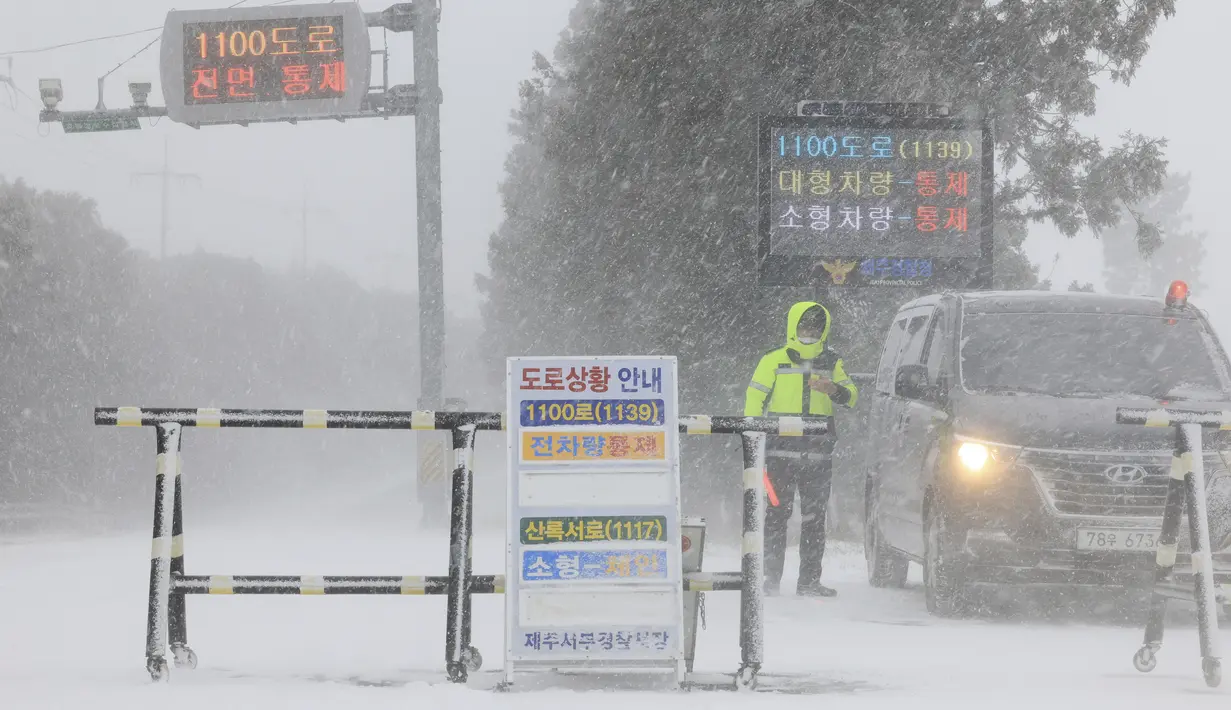 Seorang petugas polisi mengontrol masuknya kendaraan di sebuah jalan di tengah hujan salju lebat di Pulau Jeju, Korea Selatan, Selasa, 24 Januari 2023. Ribuan pelancong memadati bandara kecil di pulau Jeju Korea Selatan untuk mendapatkan penerbangan setelah penundaan karena kendala angin kencang dan salju tebal.  (Park Ji-ho/Yonhap via AP)