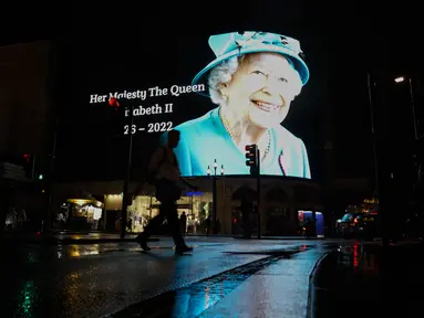 Seorang pria berjalan dengan payung melewati gambar Ratu Elizabeth II yang diproyeksikan ke layar besar di Piccadilly Circus, di London, Kamis, 8 September 2022. Ratu Elizabeth II meninggal pada hari Kamis setelah 70 tahun bertahta. Dia berusia 96 tahun. (AP Photo/Alberto Pezzali)
