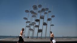 Orang-orang berjalan melewati instalasi ikonik "Umbrellas" karya pematung, pelukis, dan arsitek Yunani George Zongolopoulos, di tepi pantai Thessaloniki (23/6/2021).  Yunani bersiap menghadapi gelombang panas pertama musim panas. (AFP/ Sakis Mitrolidis)