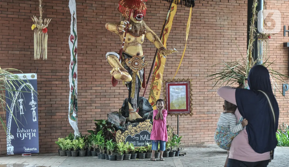 Calon penumpang KRL memfoto anaknya dengan latar belakang patung Ogoh-Ogoh yang dipajang di Stasiun Tebet, Jakarta Selatan, Selasa (1/3/2022). Patung Ogoh-Ogoh itu dipajang dalam rangka menyambut Hari Raya Nyepi Tahun Baru Saka 1944 yang jatuh pada 3 Maret mendatang. (merdeka.com/Iqbal S. Nugroho)