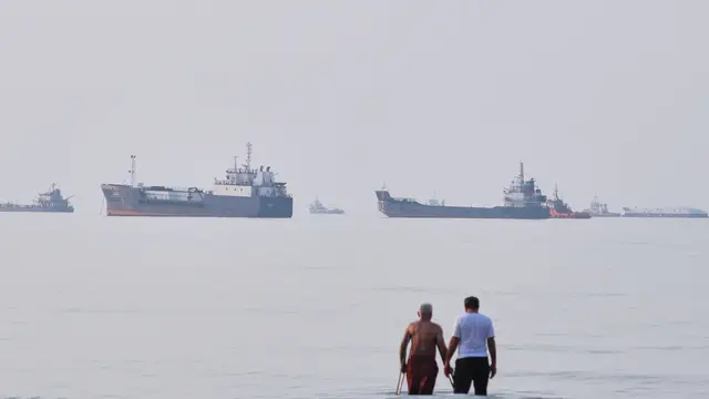 Pemandangan dua pria saat berjalan di perairan Selat Hormuz, lepas pantai Bandar Abbas, Iran, Minggu (12/7/2026). Di latar belakang, tampak sejumlah kapal tengah berlabuh. (Razieh Poudat/ISNA via AP)