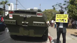 Seorang pengunjuk rasa anti-kudeta memegang poster di samping kendaraan lapis baja yang dikerahkan di luar gedung Bank Sentral Myanmar di Yangon, Senin (15/2/2021). Pasukan militer Myanmar mulai menerjunkan kendaraan lapis baja di kota-kota besar di Myanmar dan memutus akses internet. (AP Photo)