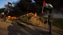 Warga Palestina membakar ban dan mengibarkan bendera nasional saat protes terhadap serangan militer Israel terhadap kamp pengungsi Jenin di perbatasan Israel-Palestina, sebelah timur Kota Gaza, 26 Januari 2023. Serangan Israel terhadap kamp pengungsi Jenin menjadi salah satu pertempuran paling mematikan dalam beberapa tahun terakhir. (AP Photo/Fatima Shbair)