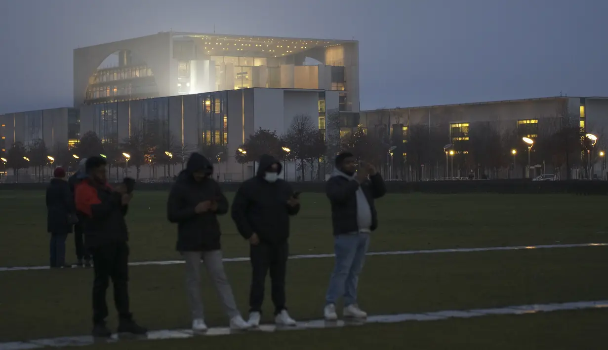 Turis berdiri di depan kanselir yang diterangi cahaya di Berlin, Jerman, Rabu (29/12/2021). Pembatasan yang berlaku selama beberapa hari terakhir termasuk membatasi pertemuan pribadi hingga 10 orang. (AP Photo/Markus Schreiber)