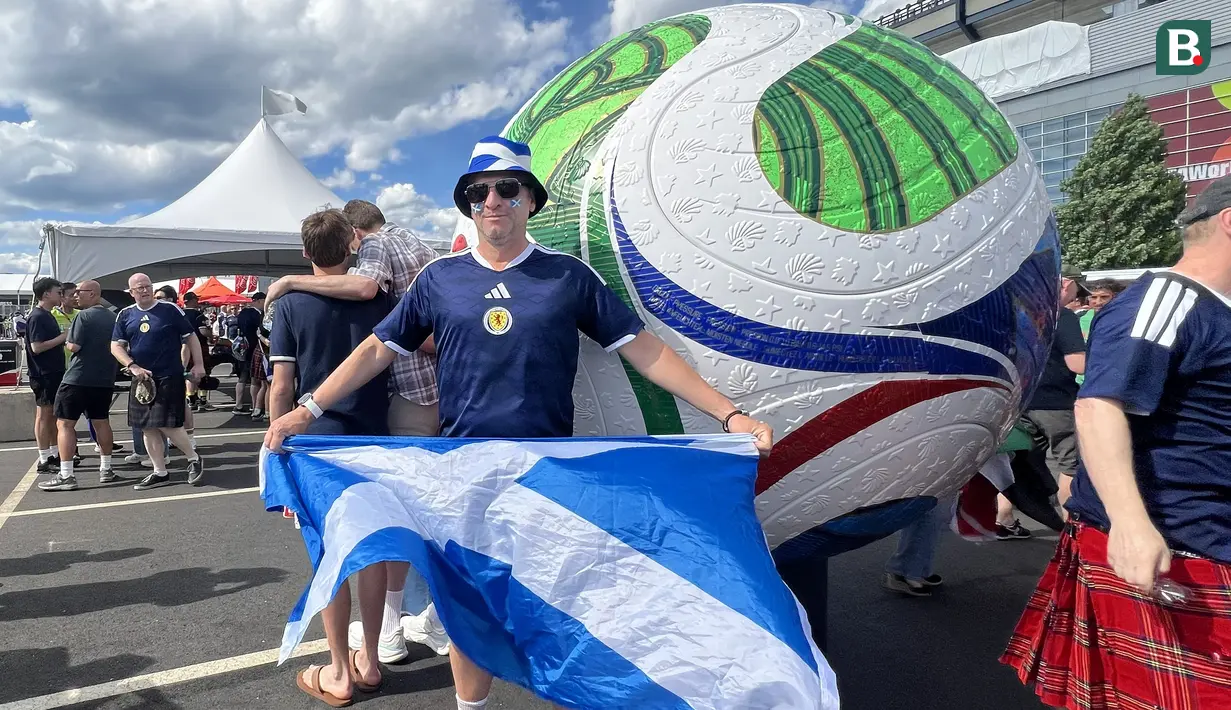 Suporter Skotlandia berpose sambil membentangkan bendera Saltire di depan replika bola resmi Piala Dunia 2026 di area fanzone Gillette Stadium, Foxborough, Amerika Serikat, Jumat (19/6/2026), menjelang laga melawan Maroko. Beragam atribut khas yang dikenakan Tartan Army turut menyemarakkan atmosfer Piala Dunia 2026. (KLY Sports/Hery Kurniawan)