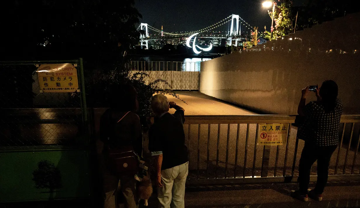 Warga melihat simbol Paralimpiade yang menyala dengan latar belakang Jembatan Pelangi dan Menara Tokyo di tepi Pantai Odaiba, Tokyo, Jepang, 23 Agustus 2021. Paralimpiade Tokyo 2020 mulai 24 Agustus 2021 sampai 5 September 2021. (Charly TRIBALLEAU/AFP)