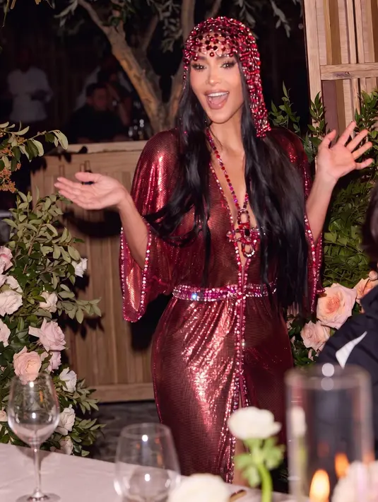 Dengan ekspresi ceria dan kedua tangan terangkat, Kim tampil semringah di meja pesta berhias bunga mawar. Kilau gaun mesh merah semakin terlihat di bawah cahaya lilin, berpadu kalung kristal dan penutup kepala senada. [@kimkardashian]