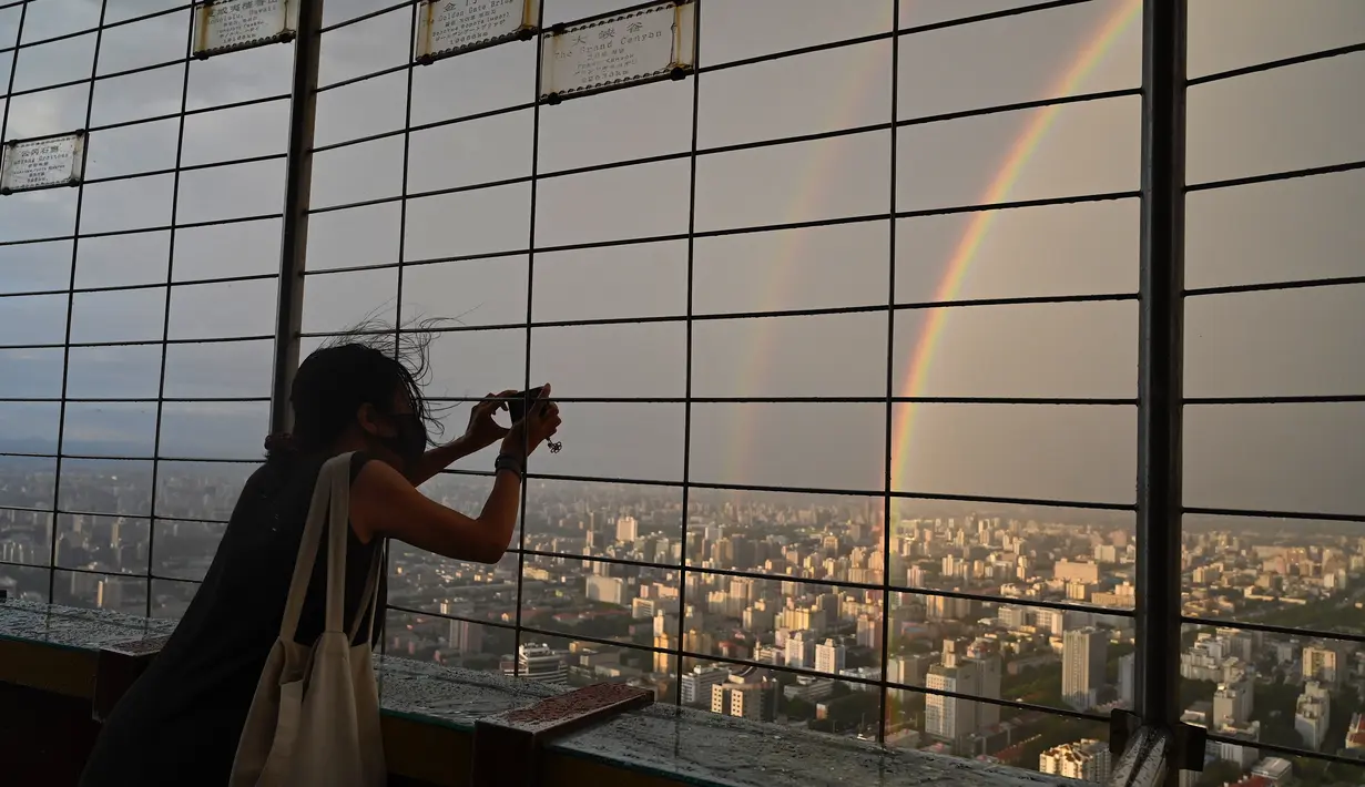 Seorang wanita mengambil foto pelangi ganda melalui pagar di Central Television Tower di Beijing (26/8/2021). Keindahan pelangi ganda dimanfaatkan warga Beijing untuk diabadikan lewat foto dan berfoto dengan latar pelangi itu. (AFP/Jade Gao)