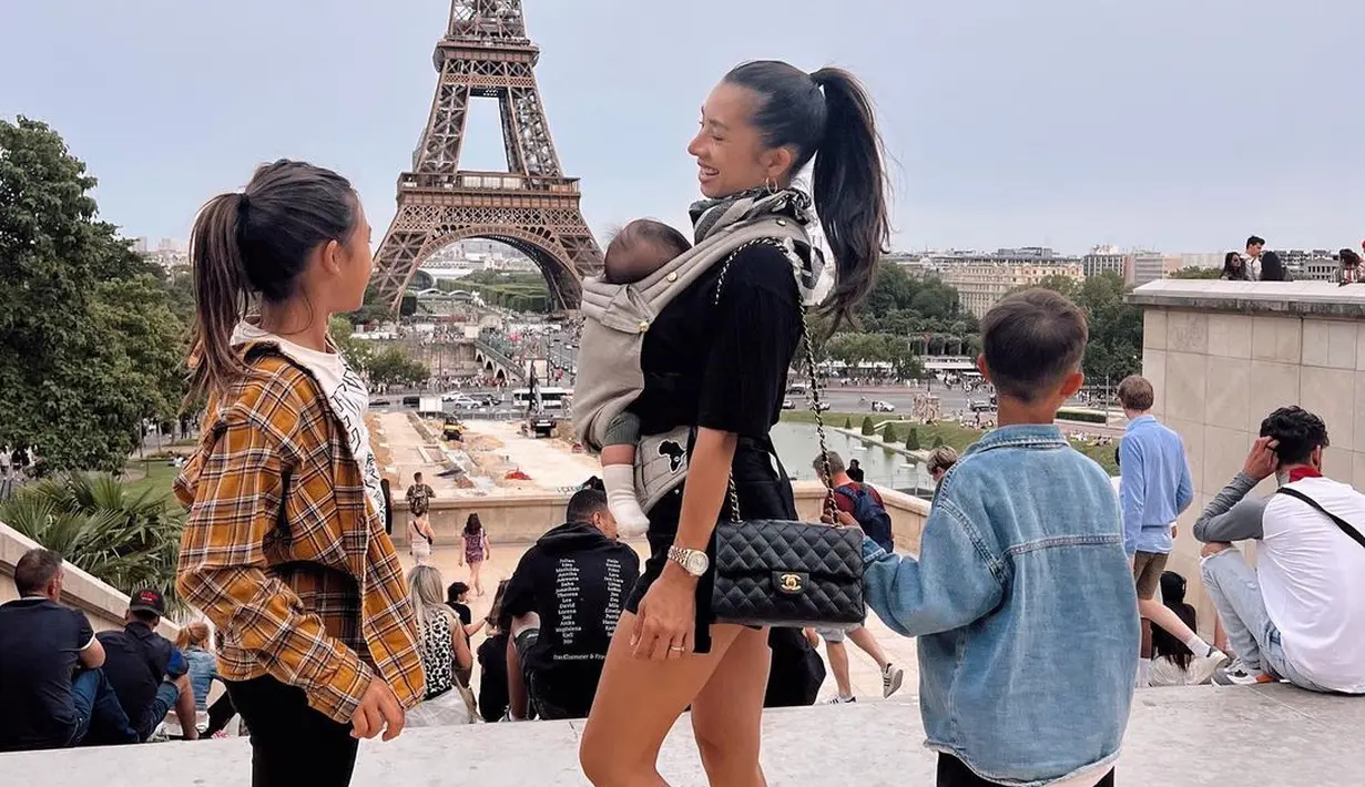 Potret gemas Jennifer Bachdim berpose dengan latar Menara Eiffel bersama buah hatinya. Ia mengenakan atasan dan celana pendek hitam, menggendong bayinya. Rambutnya dikuncir kuda tinggi dan ia menyempurnakan penampilannya dengan tas klasik Chanel, serta sneakers ungu. [Foto: Instagram/jenniferbachdim]
