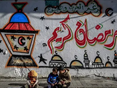 Anak-anak Palestina duduk di depan tembok dengan gambar bertema Ramadan di Kota Gaza pada 8 Maret 2024. (Foto: AFP)