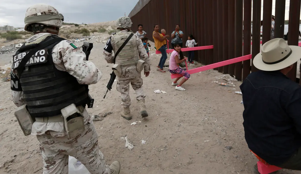 Tentara mengawasi warga dan anak-anak yang bermain jungkat-jungkit di antara pagar pemisah Meksiko dengan Amerika Serikat, Ciudad de Juarez, Meksiko, Minggu (28/7/2019). Jungkat-jungkit tersebut dirancang seorang profesor arsitektur California, Ronald Rael. (AP Photo/Christian Chavez)