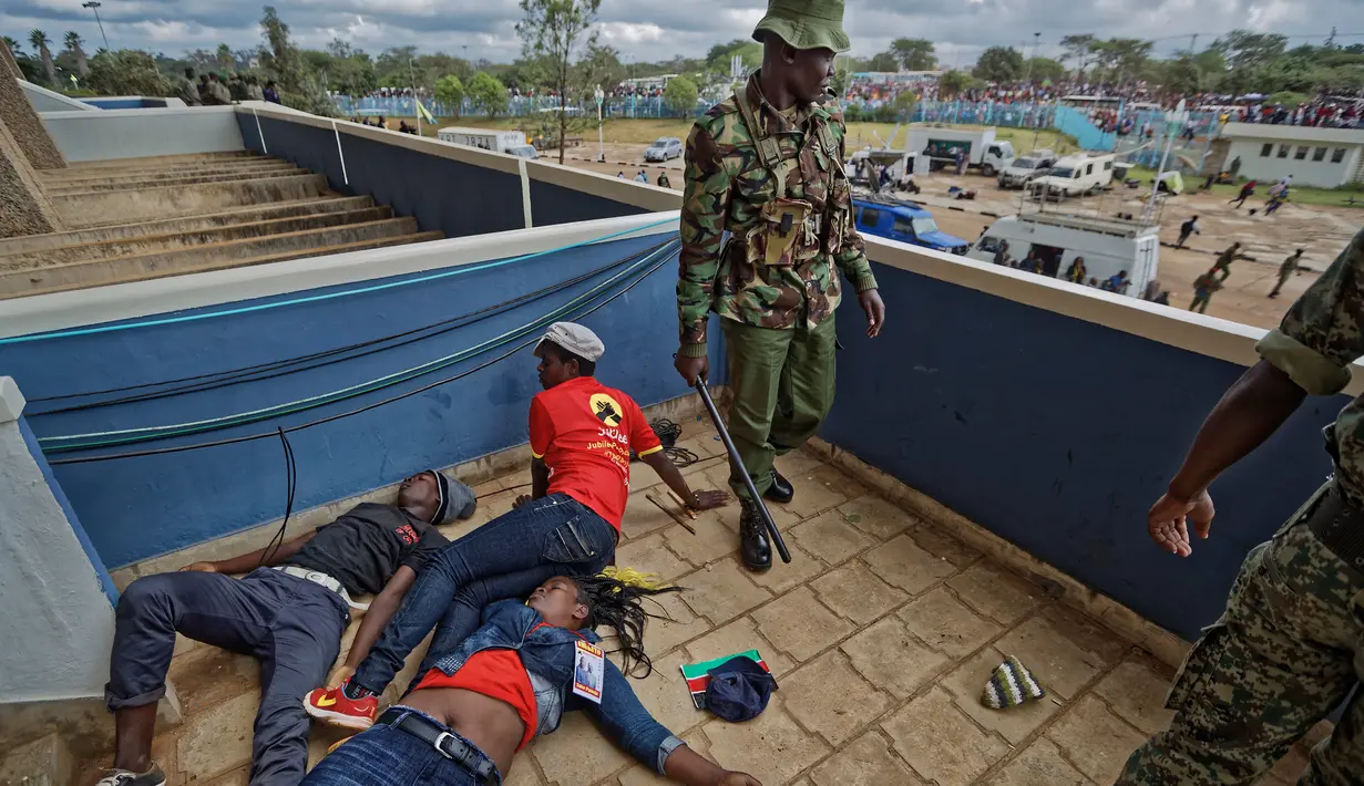 Seorang polisi berdiri di samping pendukung Presiden Kenya Uhuru Kenyatta yang terbaring tak sadarkan diri setelah dipukuli saat mereka mencoba menerobos gerbang, Nairobi, Kenya (28/11). (AP Photo / Ben Curtis)