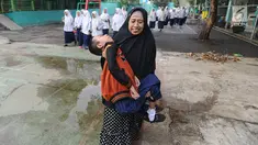 Fauzan Akmal Maulana (15), penderita Distrophia Muculor Progresive (DMP) atau penyakit otot lemah, digendong oleh ibunya, Winih Utami (49), saat bersekolah di SMP Terbuka 1 Tangerang Selatan, pada Jumat (22/3). (merdeka.com/Arie Basuki)