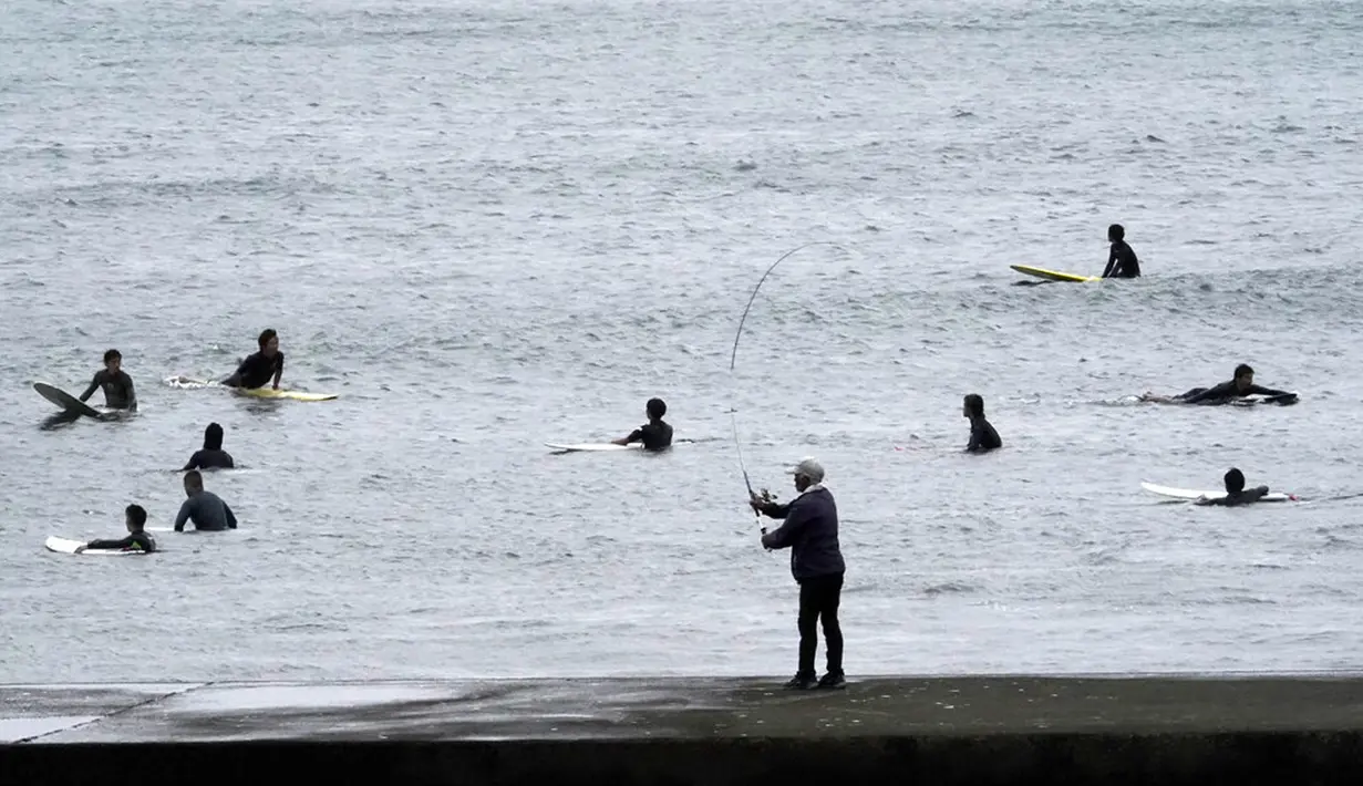 Seorang pria memancing saat peselancar berselancar di pantai Katase-kaigan yang berangin, Fujisawa, Prefektur Kanagawa, selatan Tokyo, Jepang, Kamis (24/9/2020). Badai Tropis Dolphin bergerak di sepanjang pantai Pasifik Jepang. (AP Photo/Eugene Hoshiko)