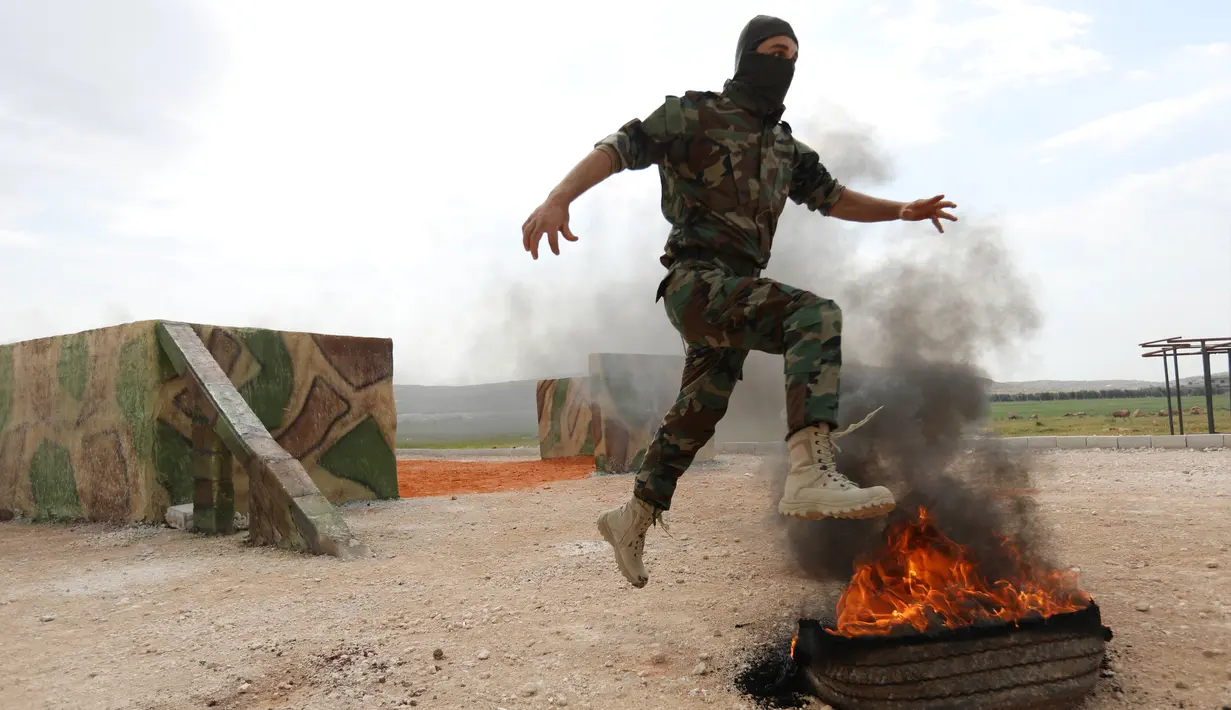 Seorang pejuang Jaysh al-Izza (the Army of Glory) melompati api saat melakukan latihan di kota Idlib, Suriah (9/4). Pejuang Jaysh al-Izza ini adalah afiliasi dari tentara Suriah yang didukung oleh Turki. (AFP/Omar Haj Kadour)