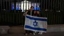 Orang-orang berdiri dengan bendera Israel di luar Gedung Putih yang menyala dengan warna biru dan putih sebagai bentuk solidaritas Amerika Serikat dengan Israel di Washington, Amerika Serikat, Senin (9/10/2023). (AP Photo/Jon Elswick)