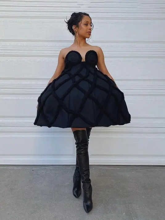 Liza Koshy mengenakan outfit dari Annaikiki yang berupa mini dress hitam mengembang di bagian bawah serta boot heels setinggi paha. (Instagram/lizakoshy).