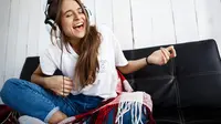 Gadis berbaju putih sedang mendengarkan musik melalui headphone dan duduk di sofa (Shutterstock/CookieStudio) 