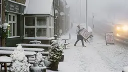 Seorang tukang daging melintasi trotoar bersalju saat Badai Eunice mendarat di Tow Law, County Durham, Inggris (18/2/2022). Jutaan warga Inggris didesak membatalkan perjalanan dan tinggal di rumah di tengah kekhawatiran gelombang tinggi, angin dan puing beterbangan. (Owen Humphreys/PA via AP)