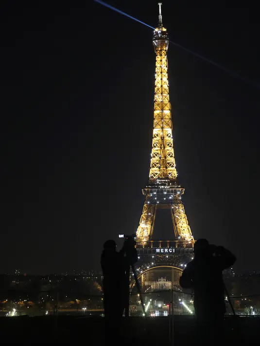 Awak media berdiri di depan Menara Eiffel dengan tulisan 'Merci" (terima kasih) yang terpampang di Paris, Jumat (27/3/2020). Menara Eiffel menampilkan pesan dukungan dan terima kasih kepada tenaga kesehatan di Prancis yang berada di garda depan memerangi pandemi COVID-19.  (AP/Thibault Camus)