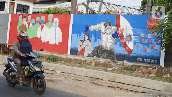 Pengendara motor melintas di depan mural bertema covid-19 di Bukit Duri, Jakarta, Minggu (30/8/2020). Mural yang dibuat petugas PPSU bertujuan mengingatkan masyarakat akan bahaya covid-19, sehubungan dengan masih tingginya jumlah kasus positif covid-19 di Jakarta. (Liputan6.com/Immanuel Antonius)