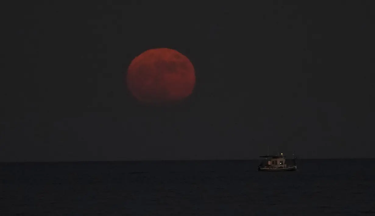 Bulan purnama terbit di atas laut Mediterania saat kapal nelayan melintas, di kota pesisir selatan Larnaca, di pulau tenggara Siprus, pada Rabu (13/7/2022). Bulan Purnama dikenal sebagai Buck Moon dan juga Supermoon. Di kondisi ini Bulan akan terlihat tampak lebih besar dan terang sehingga terasa sangat dekat dari Bumi. (AP Photo/Petros Karadjias)