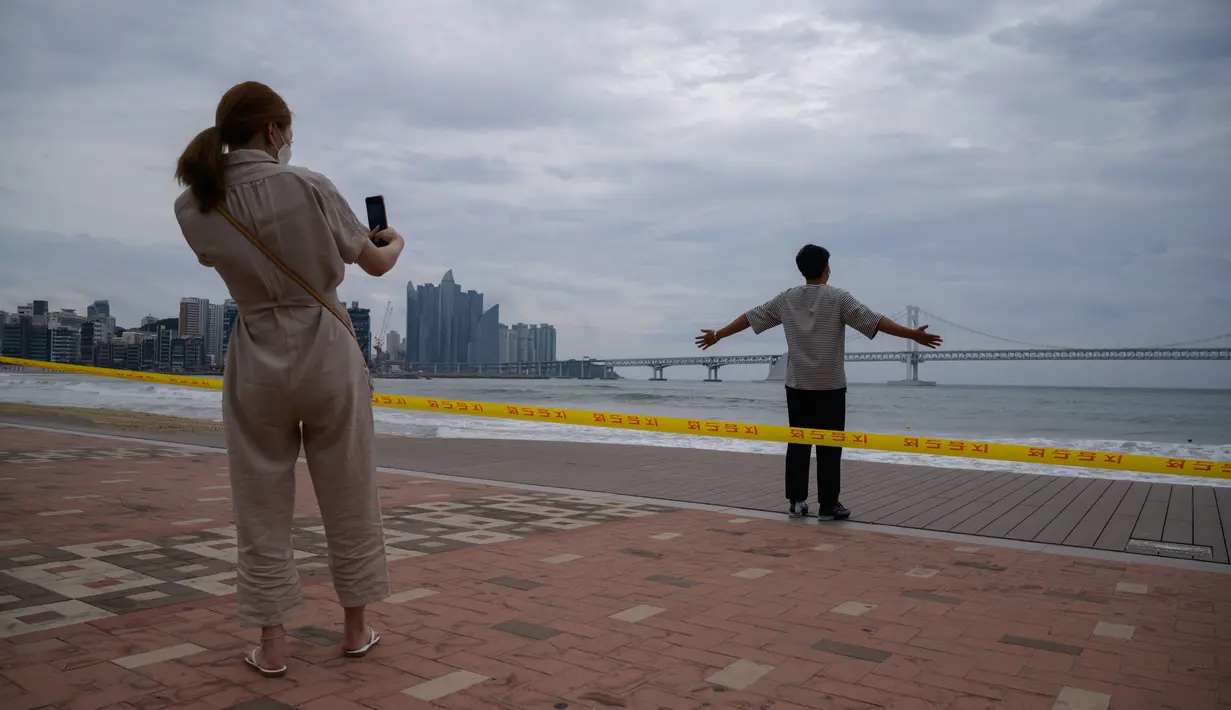 Sepasang suami istri berpose untuk foto di depan pantai yang ditutup di kawasan pejalan kaki di pantai Gwangalli di Busan (2/9/2020). Penerbangan dihentikan di Korea Selatan dan peringatan badai dikeluarkan di kedua sisi semenanjung Korea sebagai. (AFP/Ed Jones)