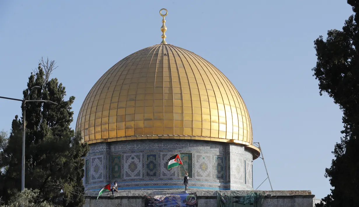 Dua warga Palestina mengibarkan bendera nasional di depan masjid Kubah Batu di kompleks masjid Al-Aqsa di Yerusalem Tua selama sholat Idul Fitri pagi, yang menandai akhir bulan suci Ramadhan, (13/5/2021). (AFP/Ahmad Gharabli)