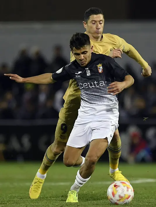 <p>Pemain AD Ceuta Aisar Ahmed (depan) berebut bola dengan pemain Barcelona Robert Lewandowski pada pertandingan sepak bola babak 16 besar Copa del Rey di Stadion Alfonso Murube, Ceuta, Spanyol, 19 Januari 2023. Barcelona mengalahkan AD Ceuta dengan skor 5-0. (JORGE GUERRERO/AFP)</p>