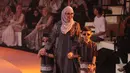 Tya Ariestya kenakan dress panjang hitam bermotif monogram dan hijab cokelat polos. Dipadukan dengan outer panjang multimotif