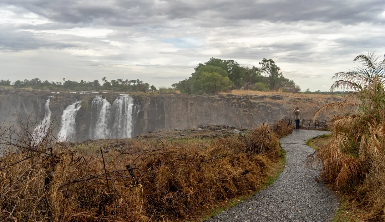 Seorang turis melihat Air Terjun Victoria yang megah di Zimbabwe (13/11/2019). Serangkaian gelombang panas telah mengeringkan sebagian besar vegetasi di sekitar situs warisan dunia UNESCO berukuran 108 meter dan lebar hampir 2 km dalam kekeringan parah. (AFP/Zinyange Auntony)