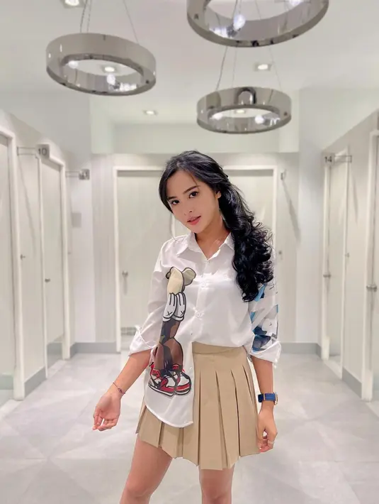 Gaya cantik Bella Bonita kenakan kemeja lengan panjang berwarna putih bermotif. Ia padukan pilihan outfitnya ini dengan mini pleated skirt cokelat muda. Foto: Instagram.