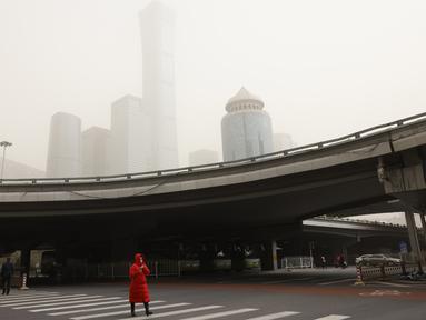 Seorang wanita menyeberang jalan dekat gedung pencakar langit saat badai pasir di Beijing (28/3/2021). Badai pasir kembali menyapu Beijing pada Minggu (28/3), mengubah langit menjadi kuning, membatasi jarak pandang dan mengganggu lalu lintas. (AP Photo/Ng Han Guan)