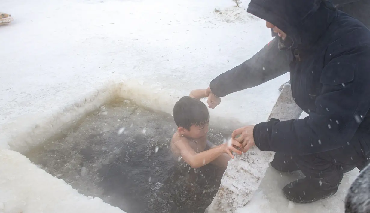 Seorang pria membantu anaknya untuk mandi di air es di Sungai Lena selama perayaan Epiphany tradisional saat suhu turun hingga sekitar -56 derajat di luar Yakutsk, Rusia (19/1/2021). Ritual ini dianggap sebagai salah satu cara untuk para umat Kristen Ortodoks. (AP Photo/Tatiana Gasich)