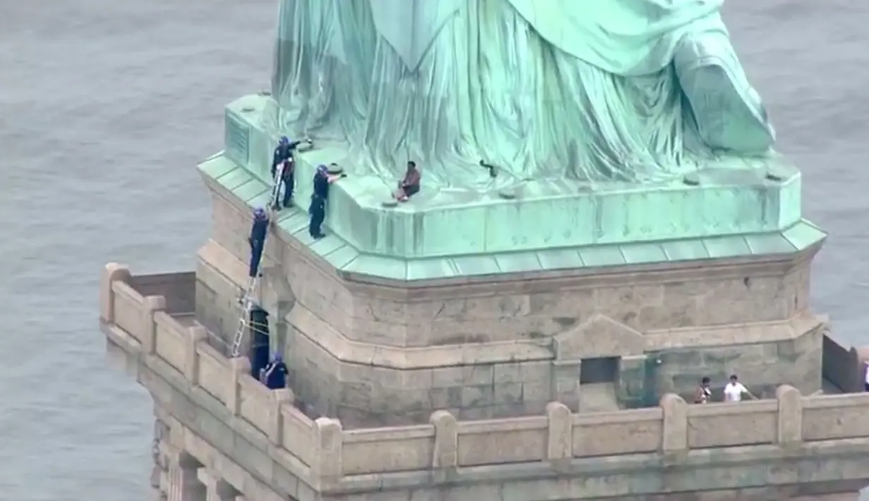 Petugas kepolisian membujuk seorang wanita yang memanjat Patung Liberty di New York, Rabu (4/7). Aksi wanita bernama Therese Okoumou ini memprotes kebijakan imigrasi pemerintah Presiden Donald Trump yang memisahkan keluarga migran. (AFP/PIX11 News/HO)