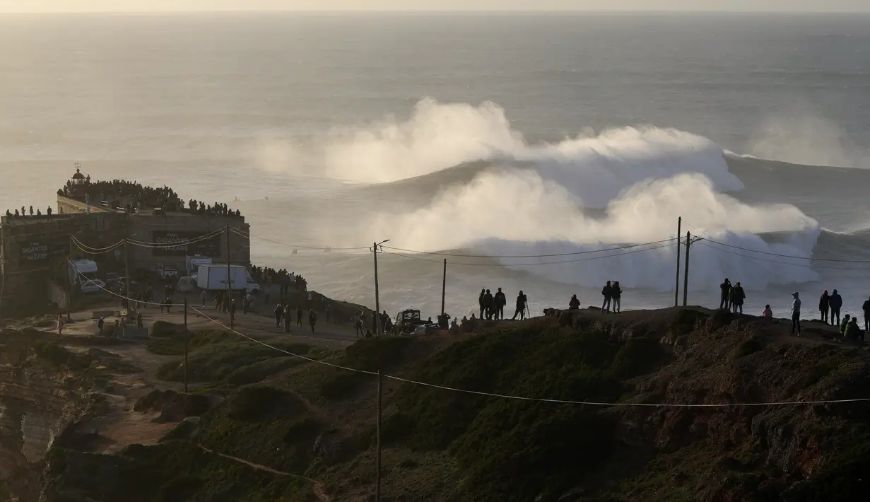 Orang-orang berdiri di atas tebing dan dekat mercusuar Nazare untuk menyaksikan sesi selancar ombak besar di Praia do Norte, Portugal, Jumat (7/1/2022). Setiap tahunnya peselancar profesional menantang adrenalinnya mengarungi ombak besar dan tinggi di Pantai Praia do Norte. (AP Photo/Armando Franca)