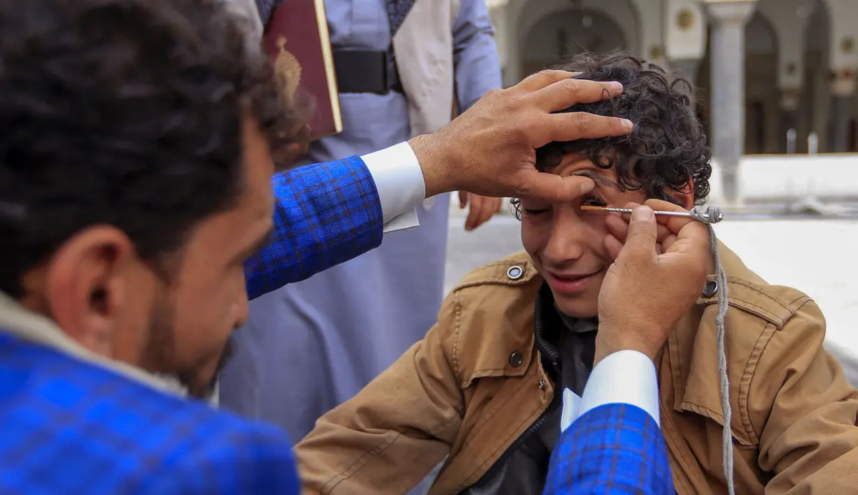 Seorang pria Yaman mengoleskan kosmetik tradisional Kohl ke kelopak mata remaja selama bulan puasa Ramadhan di Masjid Sanaa (16/4/2021).  Di Yaman, salah satu tradisi khas menandai Ramadhan yang masih lestari adalah aksi para pemuda yang merias mata dengan kohl. (AFP/ Mohammed Huwais)