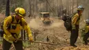 Sebuah buldoser melewati petugas pemadam kebakaran yang memotong vegetasi untuk memperluas jalur api di Oak Fire dekat Mariposa, California, Amerika Serikat, 25 Juli 2022. Petugas pemadam kebakaran terus berjuang melawan kebakaran hutan terbesar di California. (DAVID MCNEW/AFP)
