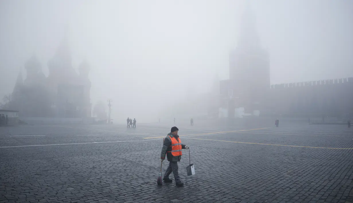 Seorang pekerja kota berjalan di Lapangan Merah melewati Menara Spasskaya dan Katedral St. Basil terlihat melalui kabut di Moskow, Rusia, Selasa (2/11/2021).  Kabut tebal menyelimuti ibu kota Rusia pada Selasa, menyebabkan banyak penundaan penerbangan di bandara Moskow. (AP Photo/Pavel Golovkin)