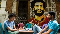 Dua pemuda duduk bersantai dengan latar belakang mural bintang Liverpool, Mohamed Salah di sebuah kedai kopi di pusat kota Kairo, 30 April 2018. Mural pemain timnas Mesir tersebut menghiasi sudut negara asalnya. (AFP Photo/KHALED DESOUKI)