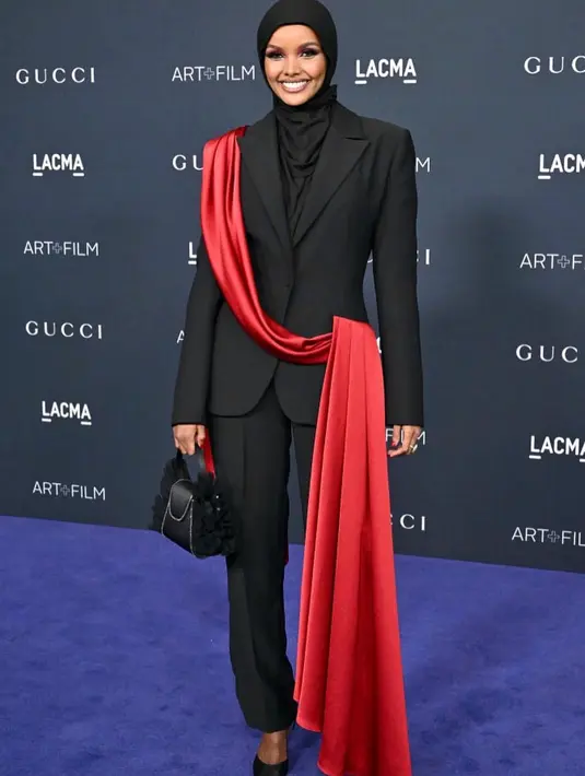 Kali ini, Halima Aden memilih tampilan elegan. Dengan set blazer dan celana panjang hitam, Halima memilih detail unik di bagian blazernya, yaitu selipan selendang merah panjang. [Foto: Instagram/halima]