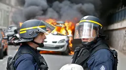 Dua petugas kepolisian berada di dekat mobil dinas yang terbakar akibat demo buru di Paris, Prancis (18/5). Demo buruh tersebut menentang kekerasan polisi dan melawan reformasi hukum yang berakhir bentrok dengan polisi. (REUTERS / Charles Platiau)