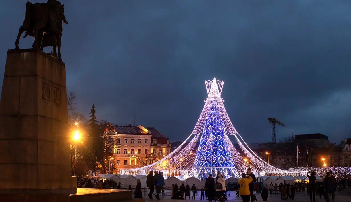 Orang-orang berjalan di dekat pohon Natal raksasa yang menerangi Alun-Alun Katedral di Vilnius, Lithuania, Minggu (1/12/2019). Secara tradisi, pohon Natal Vilnius biasanya berupa rangka logam yang ditutupi dengan dekorasi dan cabang-cabang pohon dari hutan. (AP/Mindaugas Kulbis)