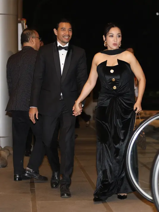 Hesti memilih strapless dress hitam dengan tambahan detail selendang di kerah, menciptakan siluet dramatis yang tetap anggun. Edi Borne hadir mendampingi dengan setelan jas hitam yang solid dan rapi, menjadi pasangan yang tampil stunning tanpa usaha berlebihan. [Dok/KLY/Budy Santoso].