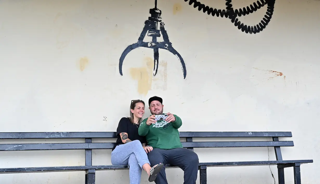 Pasangan mengambil foto selfie di bawah karya seni grafiti dari pencapit mesin permainan hiburan, yang menyandang ciri khas seniman jalanan Banksy, di dinding di Gorleston-on-Sea, pantai Timur Inggris pada 8 Agustus 2021. Karya tersebut belum diklaim oleh Banksy. (JUSTIN TALLIS / AFP)