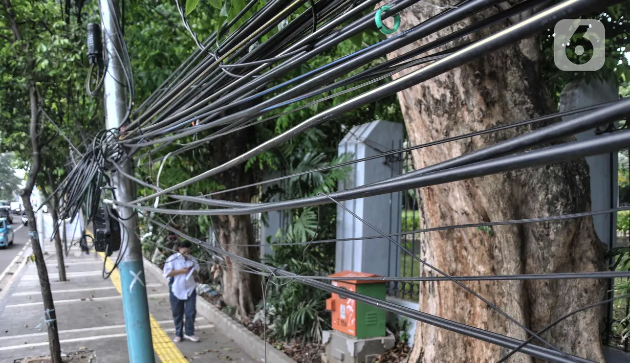 Pejalan kaki melintas di bawah jaringan kabel utilitas yang menjuntai di Jalan Proklamasi, Pegangsaan, Kecamatan Menteng, Jakarta Pusat, Kamis (6/10/2022). Minimnya pengawasan dalam tahap penyelesaian proyek revitalisasi trotoar oleh pihak terkait menyebabkan jaringan kabel utilitas di kawasan tersebut semrawut  yang dapat membahayakan keselamatan pengguna jalan, terutama saat musim penghujan. (merdeka.com/Iqbal S. Nugroho)